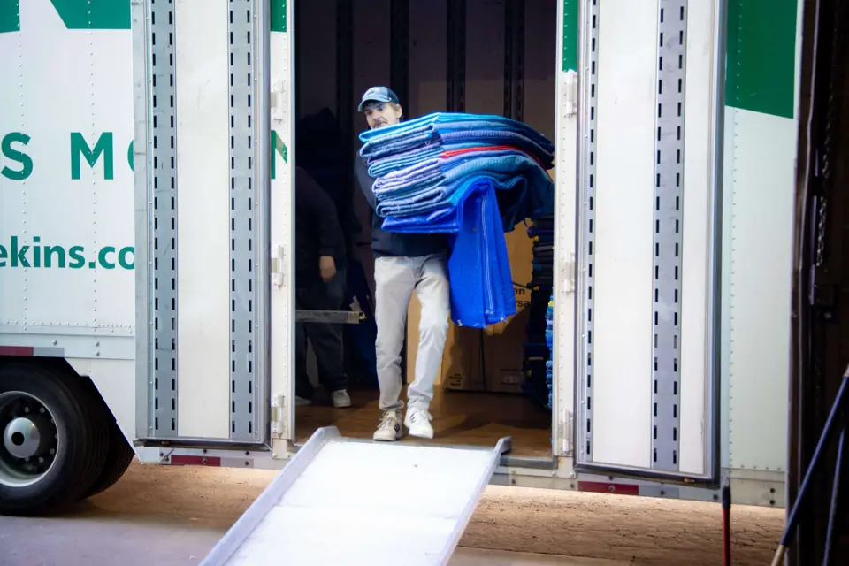 An O'Brien's mover carrying a stack of blue furniture pads down a trailer ramp