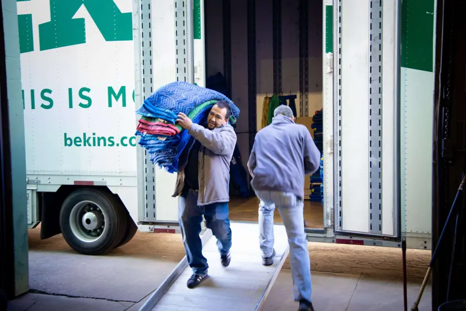Two O'Brien's movers carrying furniture pads down the ramp of a Bekins trailer
