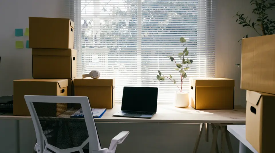 Office desk with a laptop surrounded by cardboard boxes in a bright room
