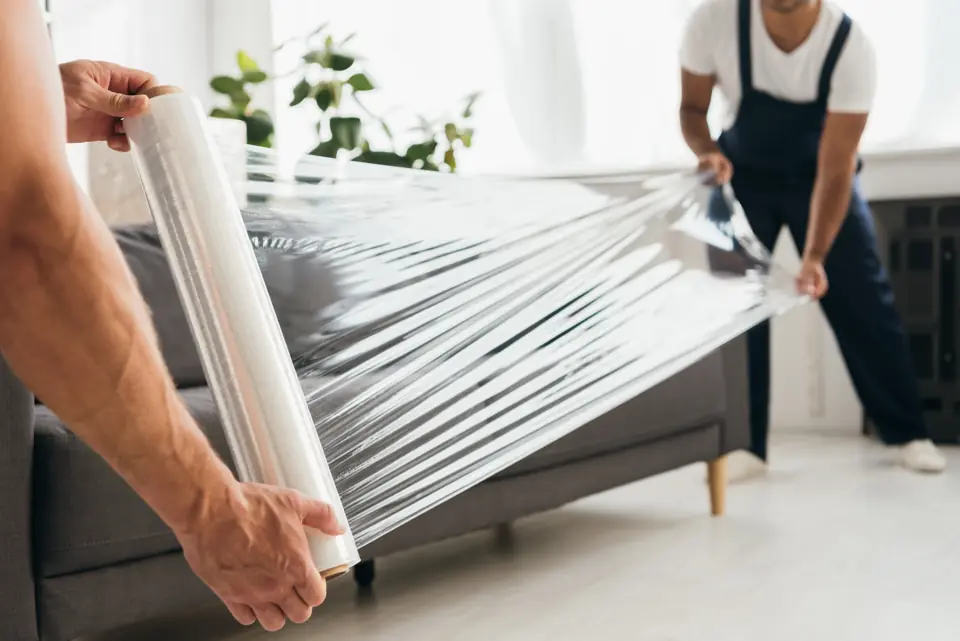 Two pairs of hands stretch-wrapping a grey sofa in plastic film, faces out of frame